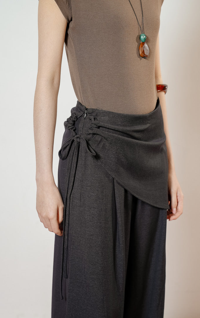 Model wanita mengenakan Nevon Side Tie Overlay Wide Leg Pants warna black, celana panjang kulot wanita berpotongan lebar warna hitam dengan detail lapisan asimetris unik di pinggang.