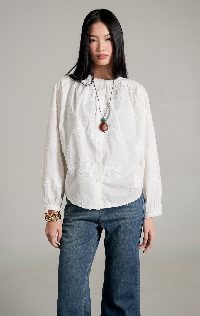 Colette Top warna putih berbahan katun dengan detail tekstur bordir bunga, dipadukan dengan celana jeans cutbray biru, kalung panjang, dan sepatu heels putih.