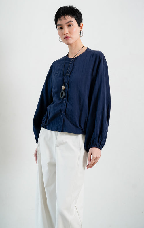 Blus wanita bahan katun ringan warna biru dongker (navy) Damian Top dengan kerah bulat (round neckline), bukaan kancing depan, lengan panjang bervolume manset karet (voluminous sleeves with elasticated cuffs), dan bagian bawah berkerut karet (elastic hem).