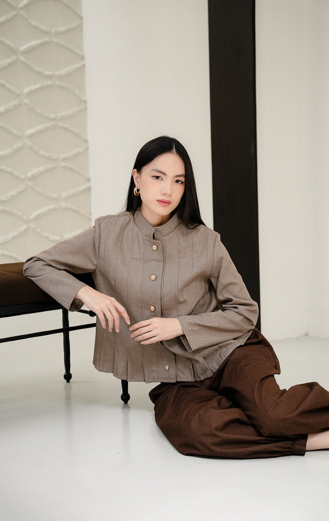 Outerwear wanita tanpa kerah (round-neck) Denali warna brown (cokelat) bertekstur lipit vertikal (pleated) dengan detail kancing emas (gold buttons) tampak depan.
