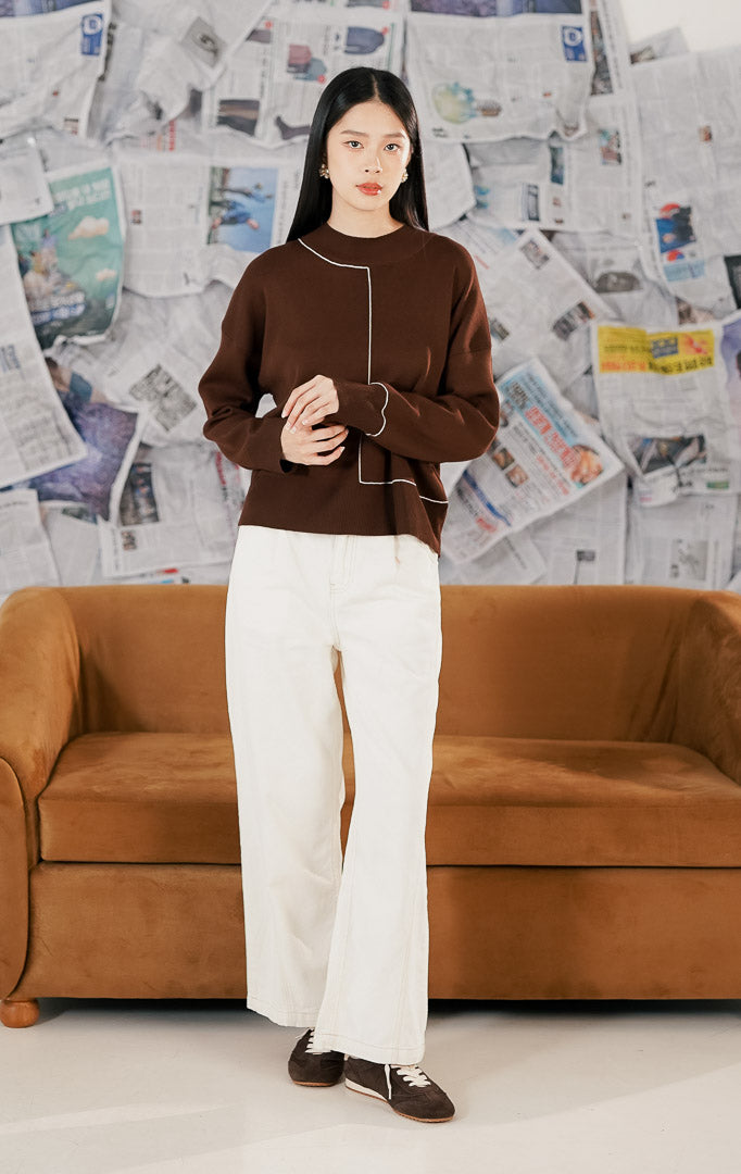 Model wanita berambut pendek mengenakan Eden Sweater Dark Brown, sebuah atasan rajut lengan panjang warna cokelat tua. Sweater memiliki detail garis putih abstrak (abstract line graphic) yang asimetris di bagian depan, dipadukan dengan celana kulot putih dan kalung mutiara.