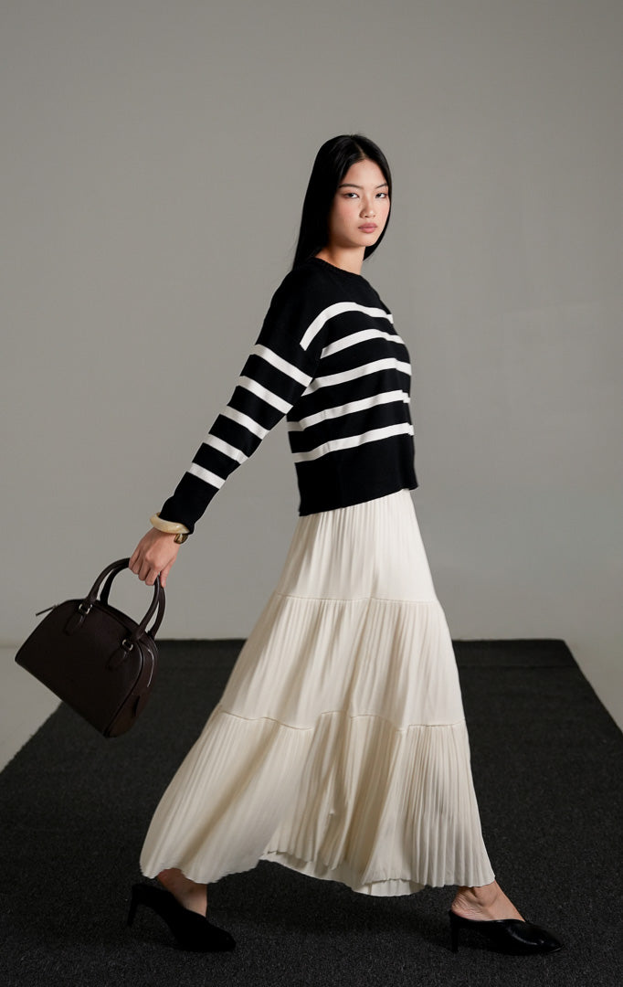 Elbie Striped Knit Sweater lengan panjang warna hitam dengan motif garis horizontal putih, dipadukan dengan rok panjang lipit warna krem dan sepatu mules hitam.