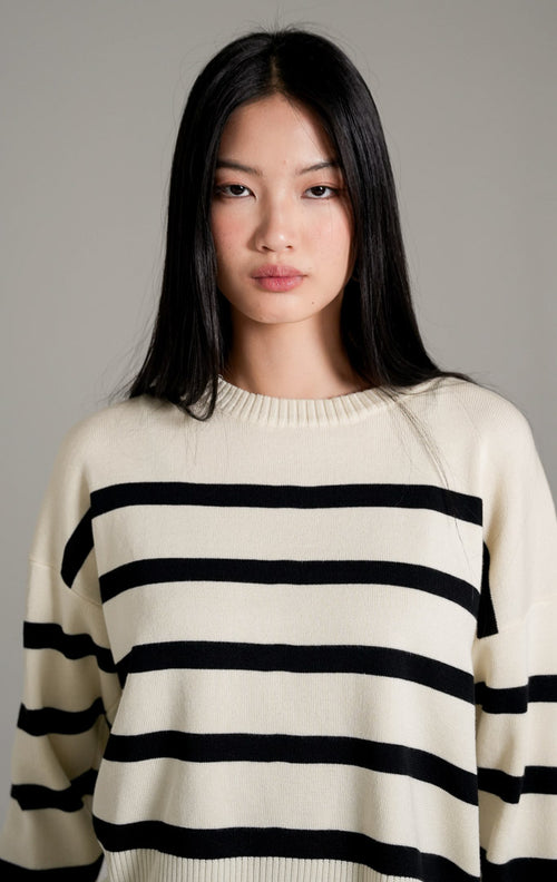 Elbie Striped Knit Sweater lengan panjang warna ivory (gading) dengan motif garis horizontal hitam, dipadukan dengan rok panjang hitam dan sandal bermotif polkadot.