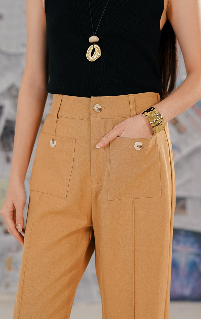 Model wanita mengenakan Elowen Pants Brown, celana panjang warna cokelat muda (camel) berbahan polyester. Celana memiliki potongan kaki lebar dengan detail jahitan vertikal di depan dan dua saku tempel besar dengan kancing dekoratif.