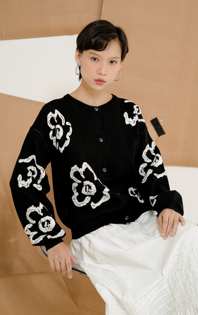 Model berpose duduk mengenakan Emmelie Flower Round-Neck Cardigan Black, produk outerwear cardigan rajut wanita lengan panjang warna hitam dengan motif bunga putih besar.