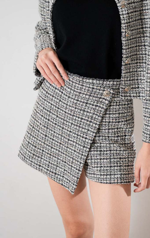 Estrella Pants Black, celana pendek bahan tweed bertekstur dengan potongan asimetris di depan dan kancing dekoratif, dipadukan dengan atasan hitam.
