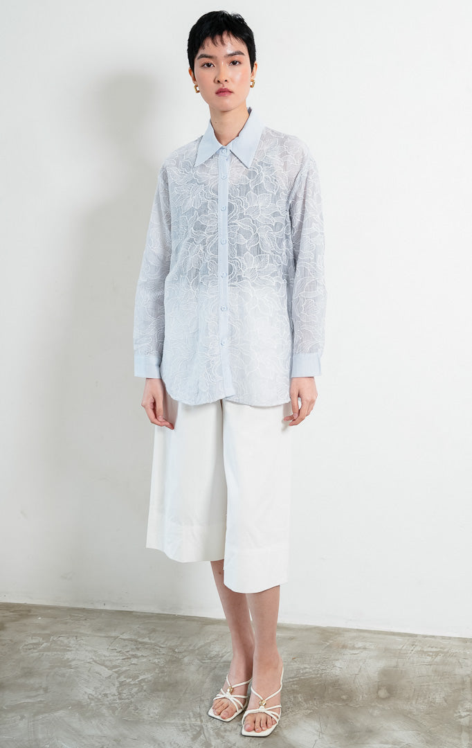 Kemeja wanita lengan panjang warna biru muda (light blue) Everlynne Top berbahan tekstur ringan (lightweight textured fabric) dengan kerah kemeja (collared neckline), bukaan kancing depan, dan detail bordir motif bunga (tonal floral embroidery).