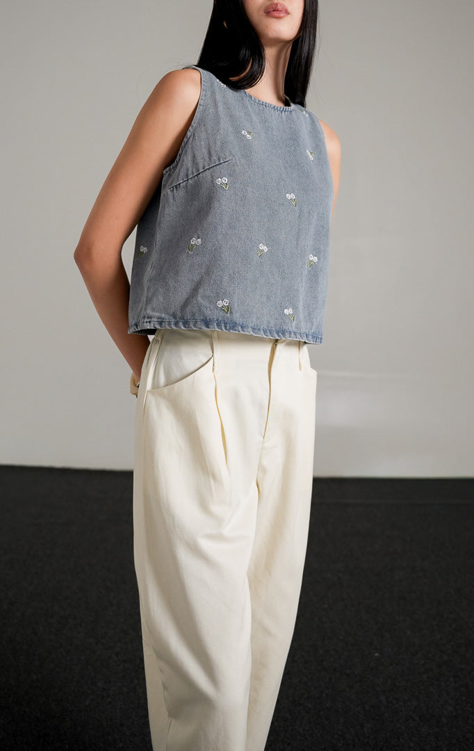 Fleura Top, atasan tanpa lengan warna light denim (biru muda) dengan detail bordir bunga kecil tersebar, dipadukan dengan celana panjang gading berpotongan longgar dan sepatu hak hitam.