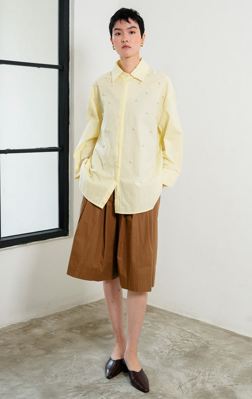Kemeja wanita lengan panjang warna kuning muda (butter yellow) Freesia Top dengan kerah klasik (classic collar), bukaan kancing depan tertutup, dan hiasan manik mutiara motif bunga (pearl bead embellishments).