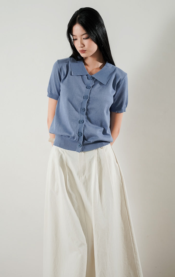Gabriela Top Dusty Blue, atasan rajut lengan pendek berkerah warna biru dusty dengan kancing depan, dipadukan dengan rok panjang putih.