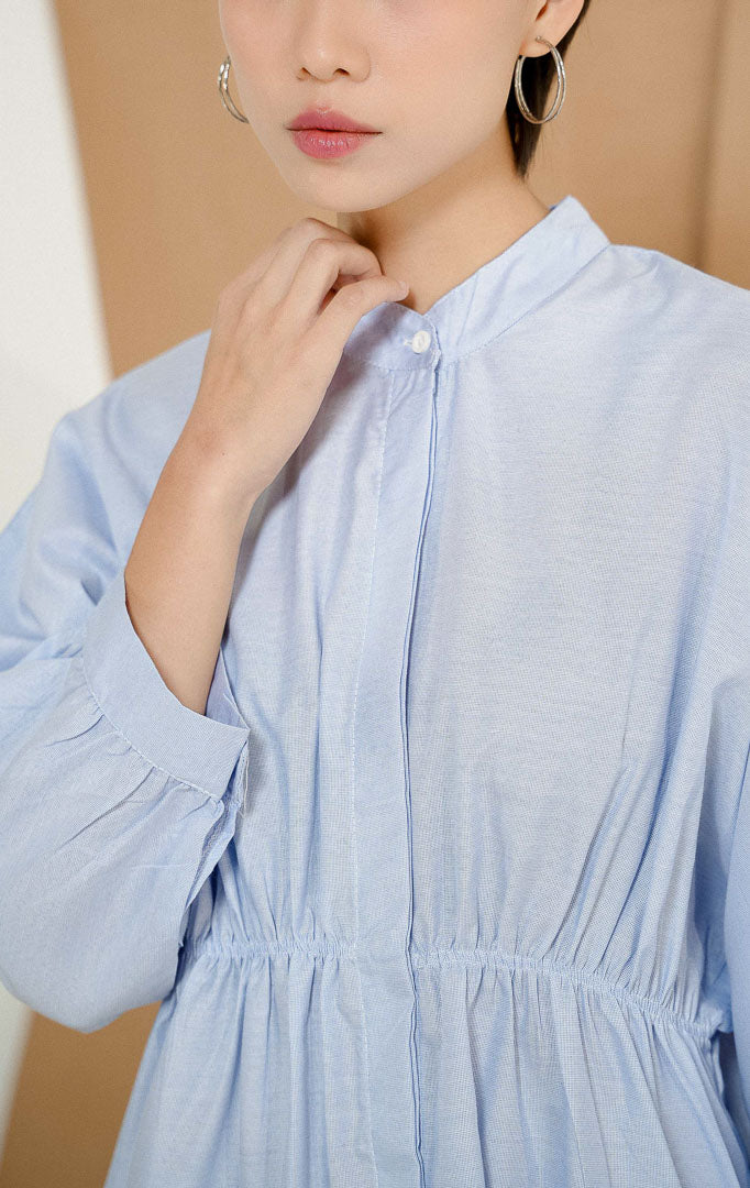 Model berdiri mengenakan Ginera Top Blue, produk atasan blouse kemeja wanita lengan panjang warna biru dengan detail kerutan di pinggang, dipadukan bersama rok panjang putih.