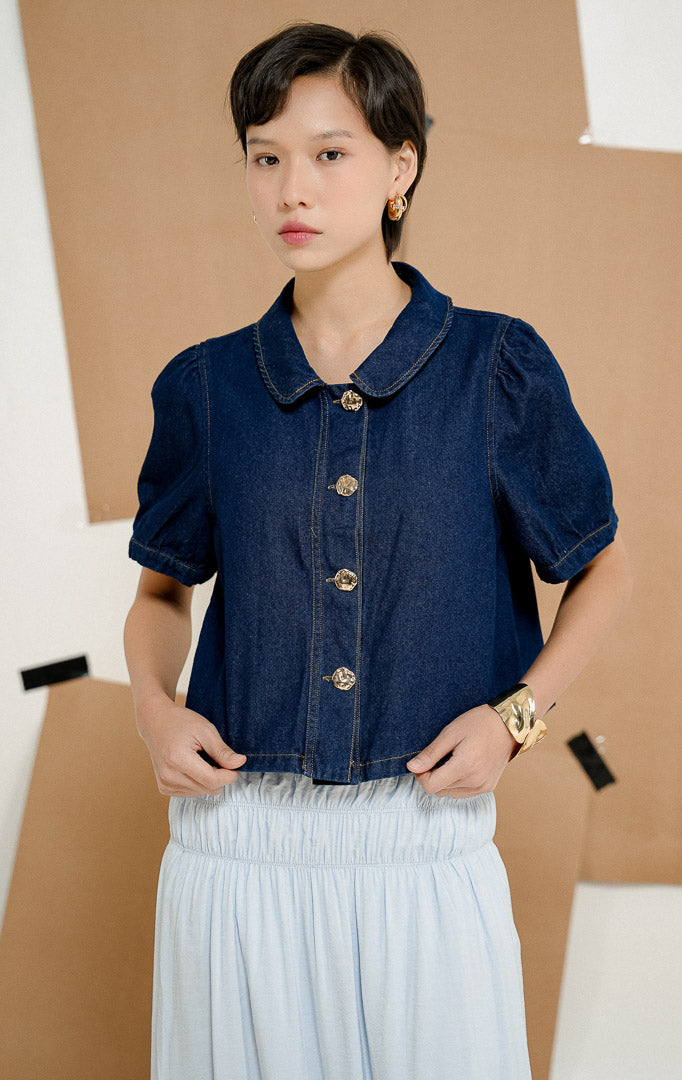Model berdiri mengenakan kemeja Hanson Peter Pan Collar Blouse in Dark Denim, produk atasan denim wanita lengan pendek dengan kerah bulat dan detail kancing emas.
