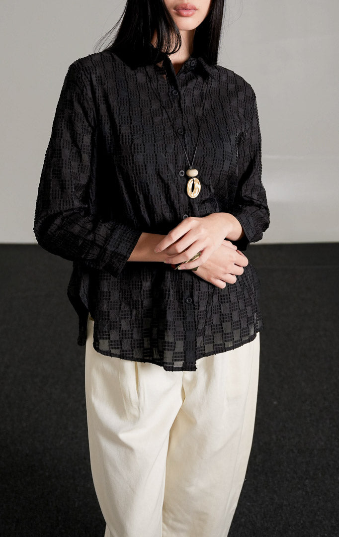 kemeja lengan panjang Hazel Top warna hitam berbahan katun dengan detail bordir pola kotak bertekstur, dipadukan dengan celana panjang broken white dan sepatu heels hitam.