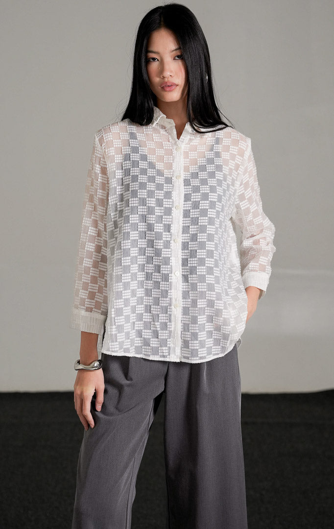 Hazel Top warna putih dengan detail bordir pola kotak semi-transparan yang memperlihatkan inner hitam di baliknya, dipadukan dengan celana kulot abu-abu dan sepatu heels putih.