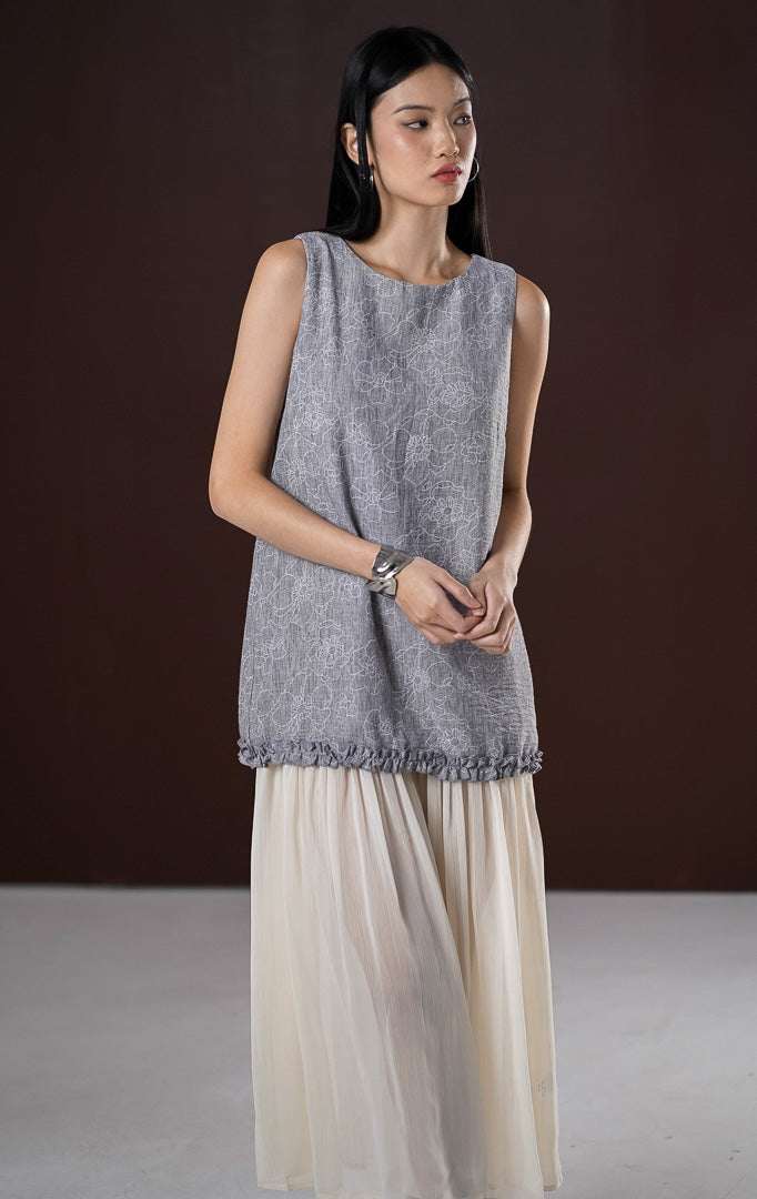 Gaun panjang wanita (maxi dress) Hermia warna abu-abu tanpa lengan (sleeveless) dengan motif floral lembut, berdetail keliman ruffle, serta tumpukan rok panjang broken white yang flowing di bagian bawah.