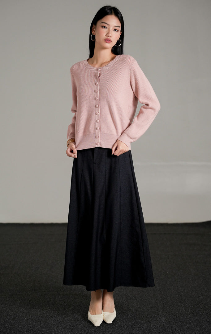 Hermione Knit Cardigan lengan panjang berwarna dusty pink dengan kancing depan tertutup, dipadukan dengan rok midi A-line hitam dan sepatu heels putih.
