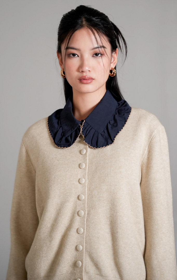 Hermione Knit Cardigan lengan panjang berwarna ivory (gading) dengan kancing depan tertutup, dipadukan dengan celana panjang bahan warna cokelat dan sepatu heels putih.