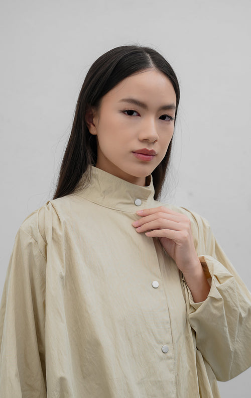 Jaket wanita warna khaki Honoria Jacket berbahan campuran crinkled cotton-nylon dengan kerah tinggi (high-neck), bukaan kancing jepret (snap-button closure), dan saku fungsional.