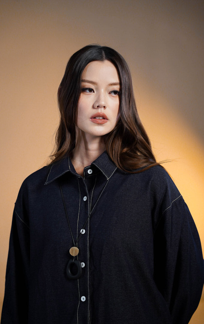 Model wanita mengenakan Howl Top Denim, kemeja lengan panjang warna biru tua (dark denim) dengan detail jahitan benang putih yang kontras di kerah dan kancing, dipadukan dengan rok panjang warna krem.