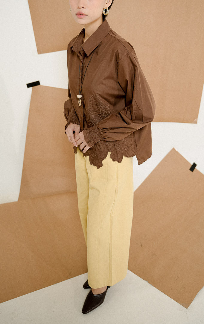 Model mengenakan Iridessa Embroidered Button-Up Shirt Dark Brown, kemeja katun wanita lengan panjang warna cokelat tua dengan detail bordir bunga dan ujung keliman bergelombang.