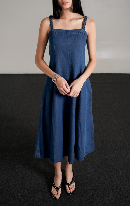 Iriska Dress berwarna indigo (biru gelap), sebuah midi dress berbahan denim tanpa lengan dengan kerah kotak, siluet A-line, dan tali bahu bertekstur, dipadukan dengan sandal hitam.