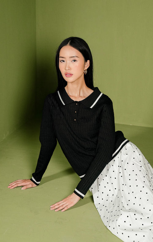 Sweater rajut wanita Isyana Sweater warna hitam lengan panjang, dilengkapi kerah polo beraksen garis putih, 3 kancing depan berwarna emas, tekstur rajut kepang (cable-knit) vertikal, dan aksen dua garis putih di ujung bawah.
