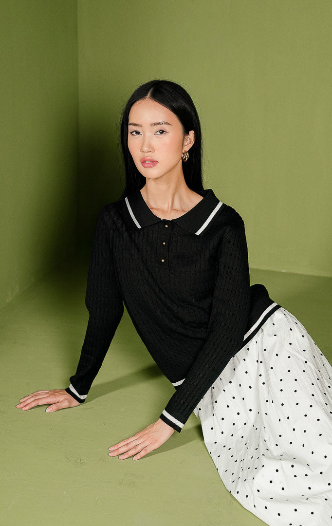 Sweater rajut wanita Isyana Sweater warna hitam lengan panjang, dilengkapi kerah polo beraksen garis putih, 3 kancing depan berwarna emas, tekstur rajut kepang (cable-knit) vertikal, dan aksen dua garis putih di ujung bawah.