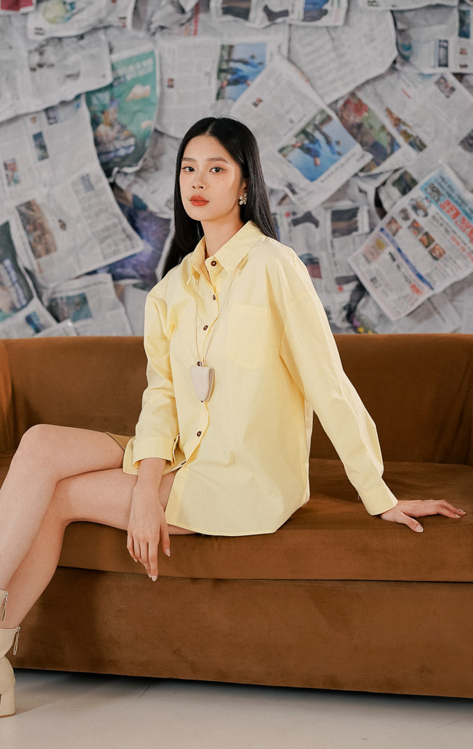 Model wanita mengenakan Izumi Top Yellow, sebuah kemeja lengan panjang model oversized warna kuning pastel (kuning muda) dengan detail kancing depan cokelat dan satu saku dada.