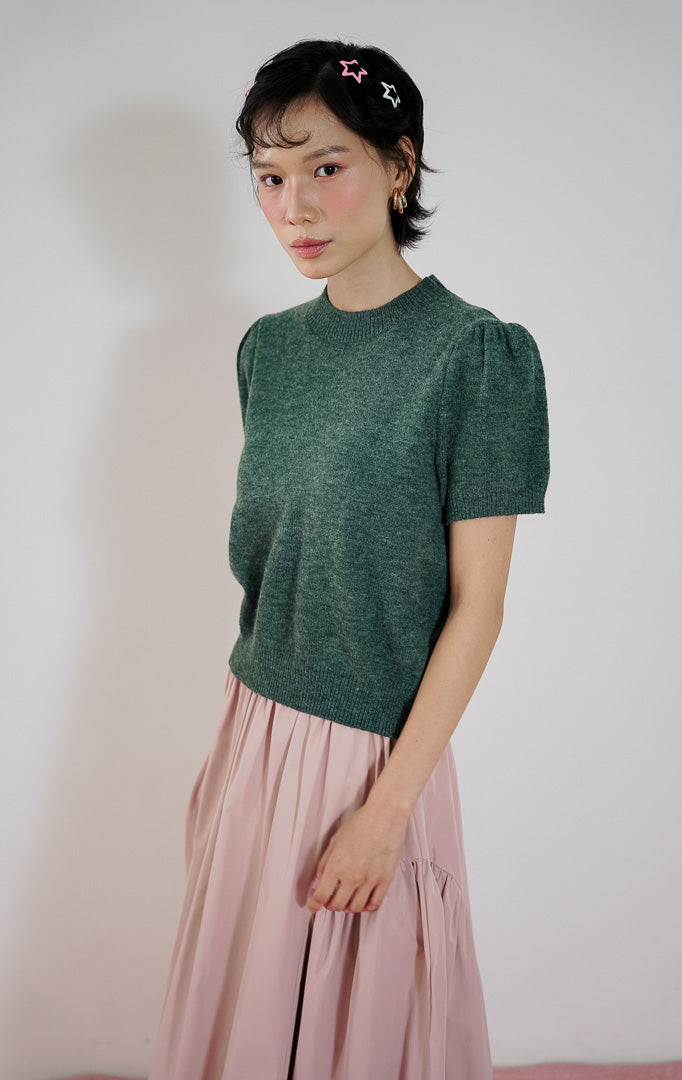 Atasan rajut wanita Jaeyun Top dari This Is April warna Dark Green dengan kerah bulat dan lengan pendek model puff (balon), dipadukan dengan rok panjang warna pink.