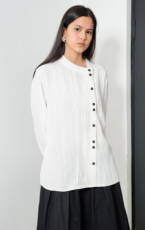 Blus wanita lengan panjang warna putih (white) Janessa Top dengan kerah asimetris (asymmetrical neckline), detail lipit pintuck vertikal di bagian depan, dan bukaan kancing tersembunyi (concealed button placket).