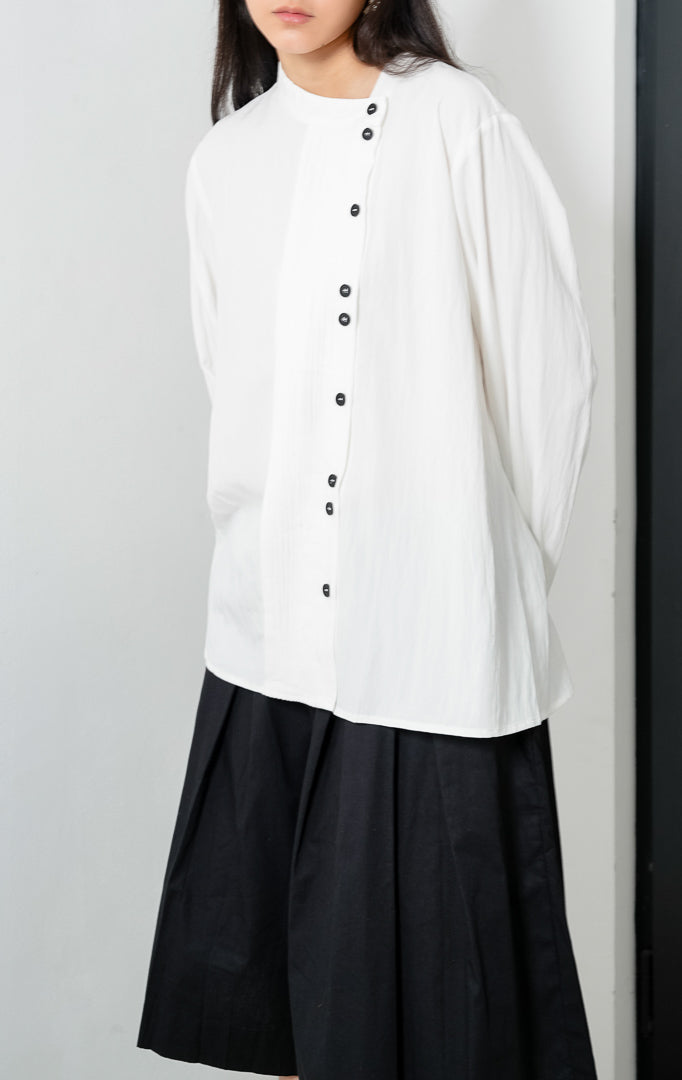 Blus wanita lengan panjang warna putih (white) Janessa Top dengan kerah asimetris (asymmetrical neckline), detail lipit pintuck vertikal di bagian depan, dan bukaan kancing tersembunyi (concealed button placket).