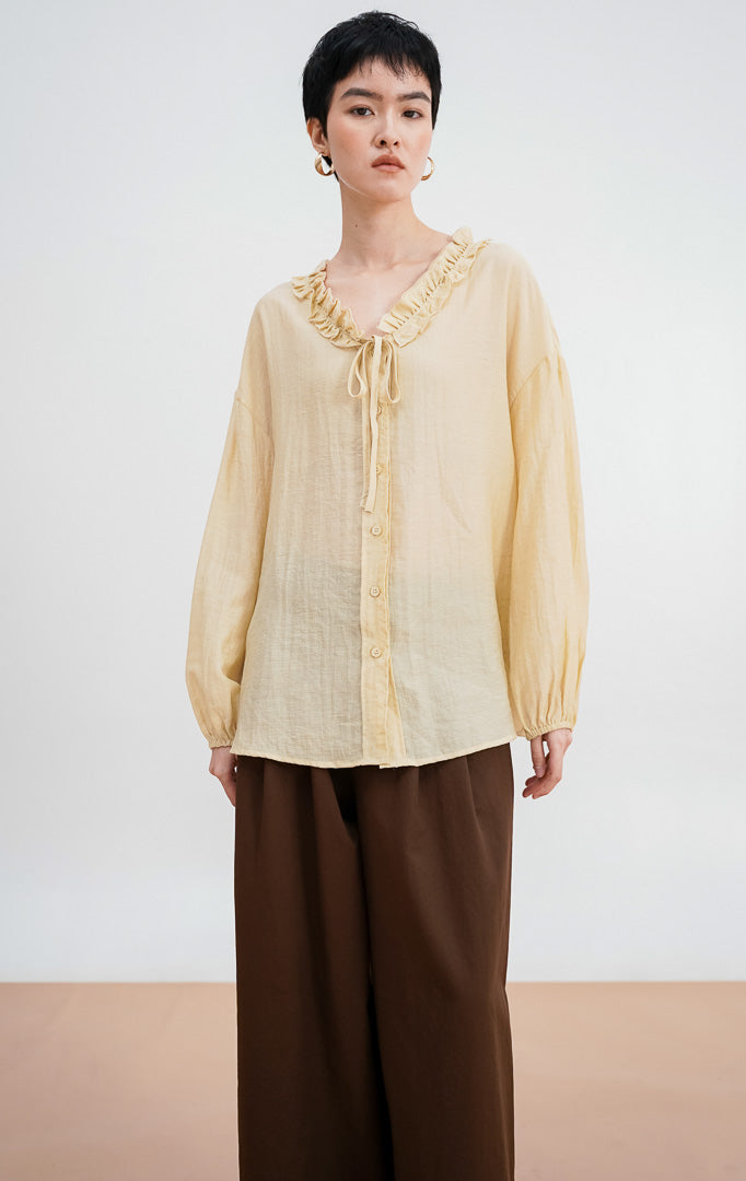 Model mengenakan Jasper Top warna ivory, atasan katun lengan panjang dengan detail kerah ruffle (kerut), tali serut di bagian leher, dan kancing depan
