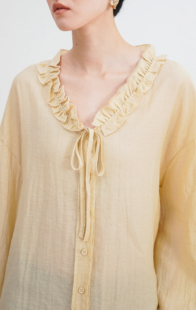 Model mengenakan Jasper Top warna ivory, atasan katun lengan panjang dengan detail kerah ruffle (kerut), tali serut di bagian leher, dan kancing depan