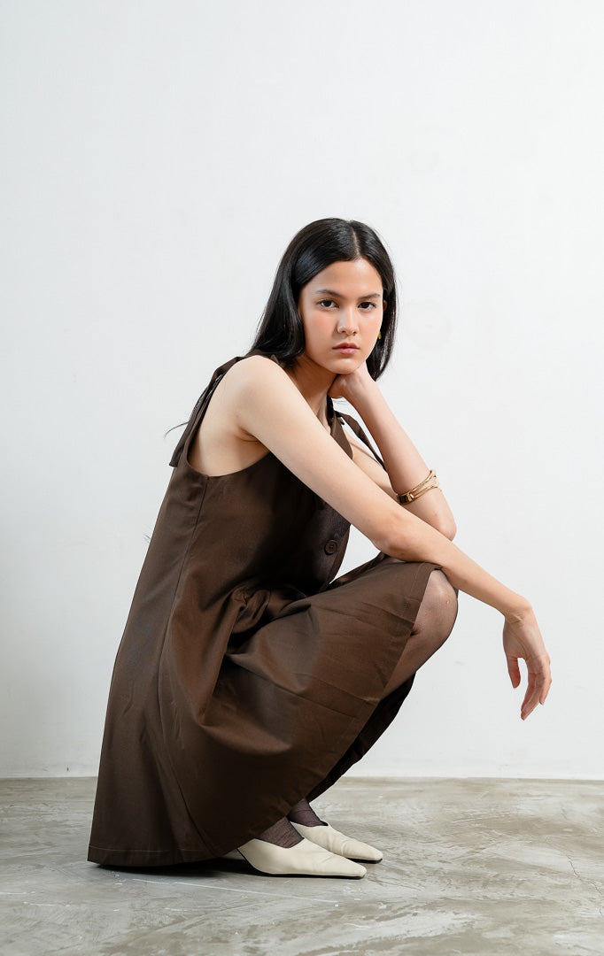 Gaun pendek wanita tanpa lengan (sleeveless mini dress) warna cokelat tua (dark brown) Joanne Dress berbahan woven halus dengan tali bahu ikat yang bisa diatur (adjustable tie shoulders), bukaan kancing depan, dan aksen kerut di panel samping.