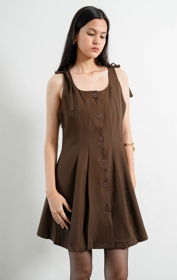 Gaun pendek wanita tanpa lengan (sleeveless mini dress) warna cokelat tua (dark brown) Joanne Dress berbahan woven halus dengan tali bahu ikat yang bisa diatur (adjustable tie shoulders), bukaan kancing depan, dan aksen kerut di panel samping.