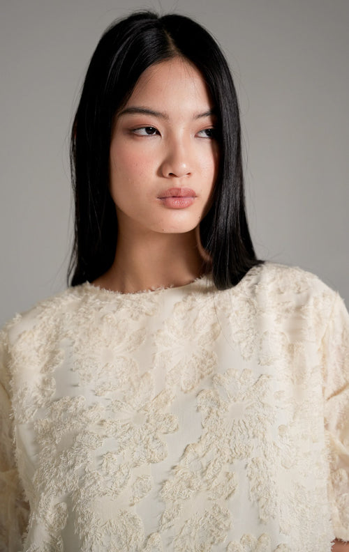 Kallani Top Ivory, atasan blus warna krem dengan tekstur fringe bulu halus dan lengan pendek semi-transparan, dipadukan dengan celana cokelat.