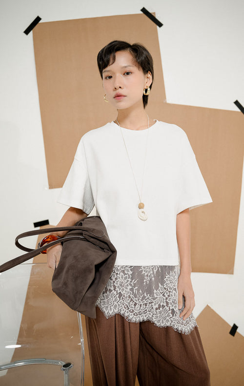 Model berdiri mengenakan Kaori Lace Hem Oversized Shirt White, produk atasan kaos kemeja wanita ukuran longgar warna putih dengan detail hiasan renda brokat abu-abu di bagian bawah.