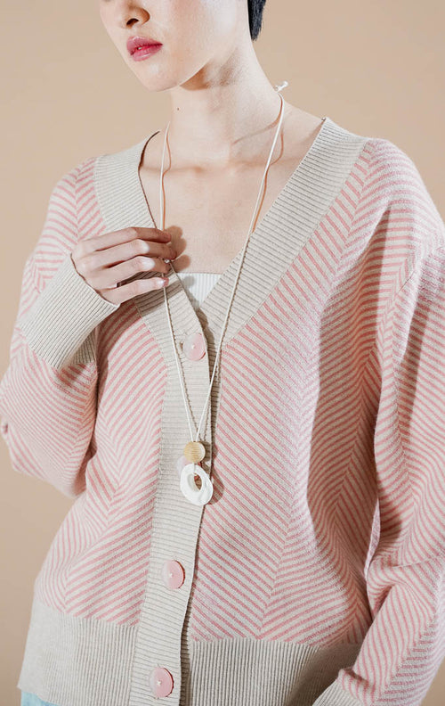 Close up model wanita mengenakan Katniss Kardigan Rajut V-neck warna pink dengan motif garis diagonal, kancing besar, dan aksesoris kalung panjang.