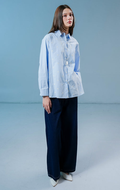 Model wanita mengenakan kemeja lengan panjang Ashford Top warna biru muda berbahan katun dengan detail kancing depan dan bordir bunga putih kecil.