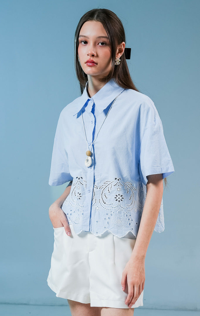 Model wanita mengenakan kemeja lengan pendek Annaleigh Top warna biru muda berbahan katun dengan detail bordir eyelet putih dan ujung bawah berpotongan scallop.
