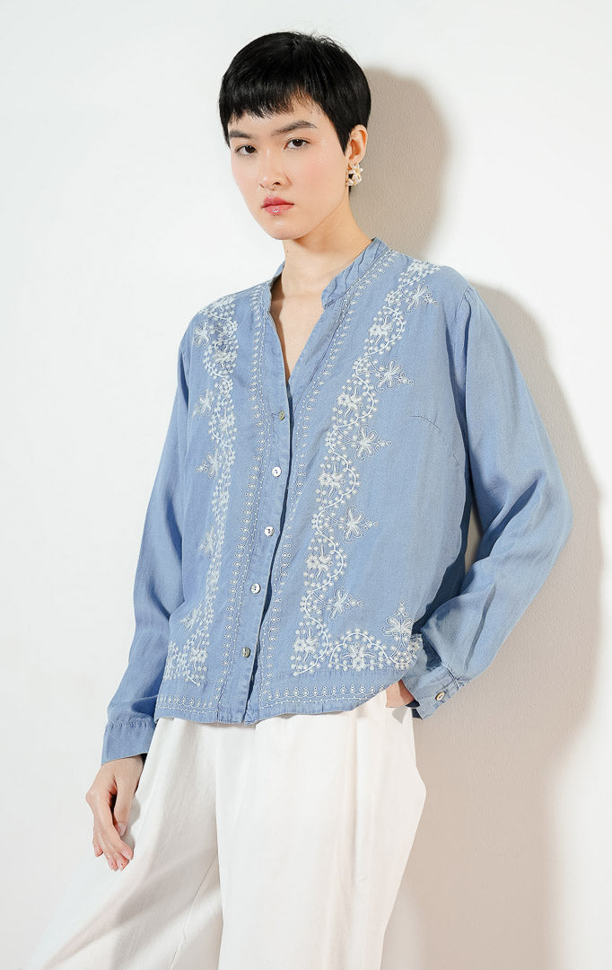 Model wanita mengenakan Brella Embroidery V-Neck Top warna blue denim, kemeja katun wanita lengan panjang berwarna biru muda dengan detail sulaman bordir bunga putih yang anggun.