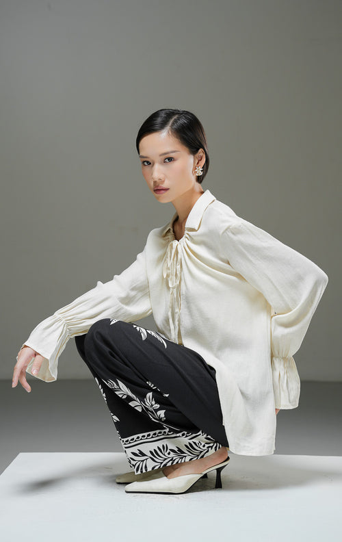 Blouse wanita Kensley Top warna ivory krem muda lengan panjang dengan detail tali pita leher (tie neck), kerah kemeja, dan kancing depan.