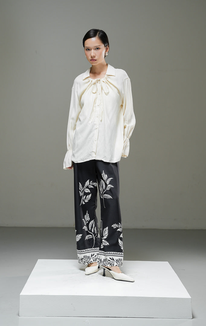 Blouse wanita Kensley Top warna ivory krem muda lengan panjang dengan detail tali pita leher (tie neck), kerah kemeja, dan kancing depan.
