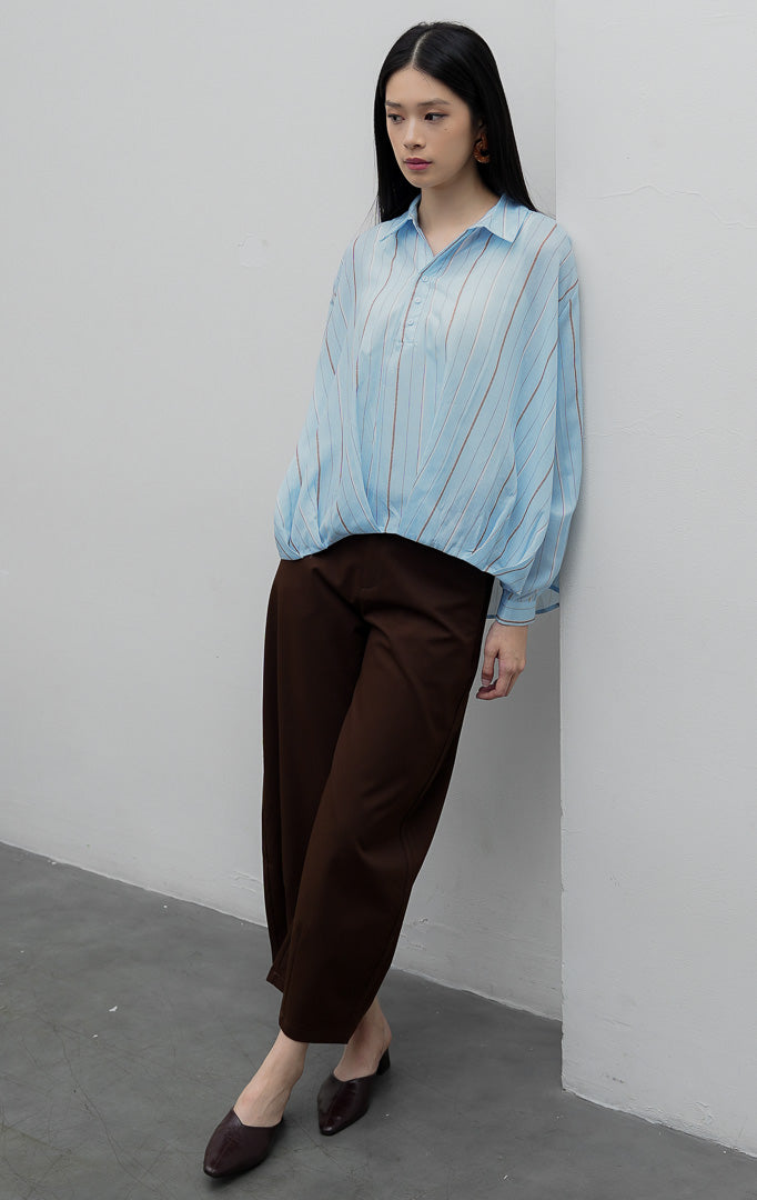 Kemeja wanita lengan panjang warna biru muda (light blue) Lorena Top dengan aksen siluet lipat silang (hi-low crossover hem), bahan katun ringan bermotif garis salur, dan bukaan kancing depan.