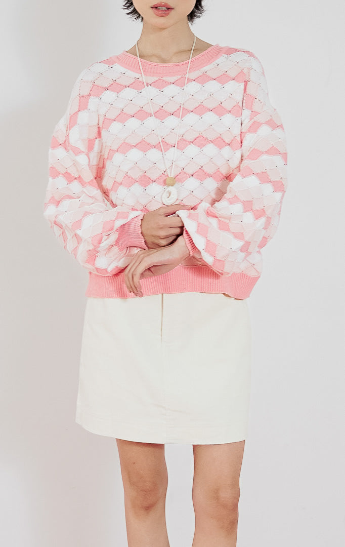 Atasan sweater rajut wanita Loryn warna pink putih dari This Is April dengan motif diamond (belah ketupat) dan kerah bulat, dipadukan dengan rok pendek putih.