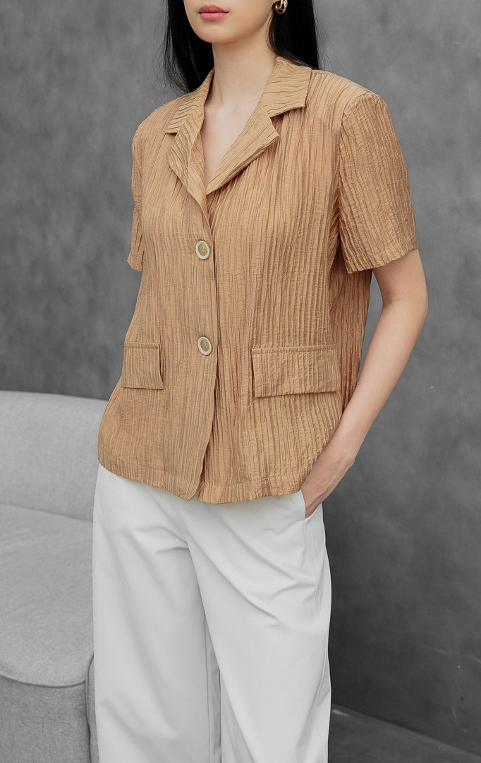 Blazer wanita lengan pendek warna cokelat muda (light brown) Lovegood Blazer berbahan crinkle polyester dengan tekstur plissé, kerah notch lapel, bukaan kancing depan, dan saku saku penutup (flap pockets).