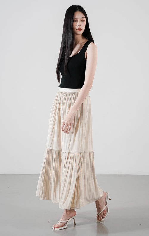 Model wanita mengenakan Lowen Skirt warna ivory, rok panjang maxi bahan velour dengan detail lipit bertingkat (tiered pleats) dan ban pinggang elastis putih.