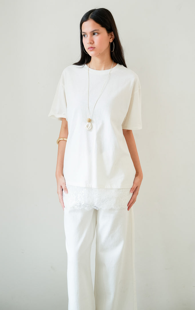 Kaos wanita lengan pendek warna putih (white T-shirt) Lyora Top dengan siluet santai (relaxed silhouette), kerah bulat, dan detail aksen hiasan renda halus di bagian kelim bawah (delicate lace hem detail).