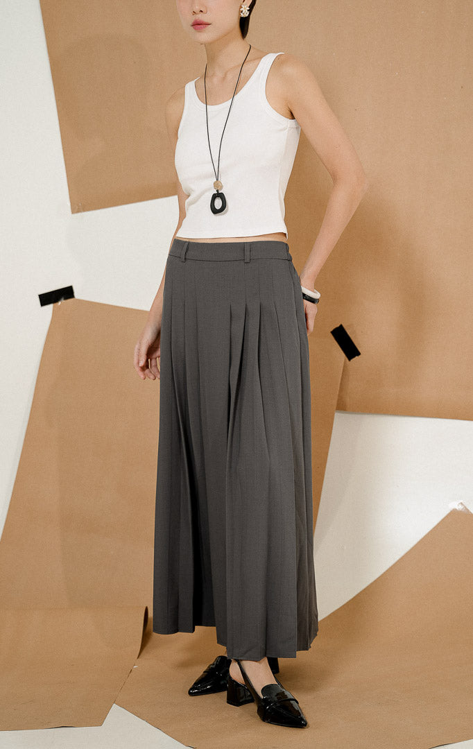 Model berdiri mengenakan Maiden Box Pleats Maxi Skirt Dark Grey, produk rok panjang wanita model maxi warna abu-abu tua dengan detail lipit kotak dipadukan tank top putih.