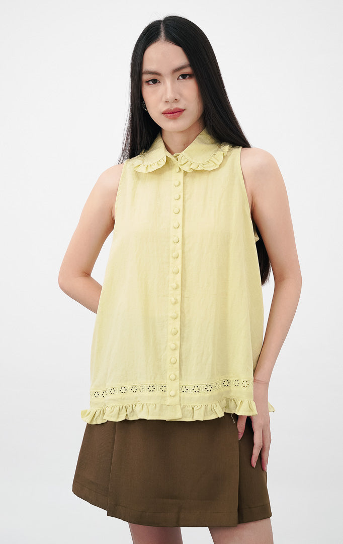 Model mengenakan Malou Top warna yellow, atasan katun tanpa lengan dengan kerah ruffle, kancing bungkus depan, serta detail bordir eyelet dan ruffle pada ujung bawah.